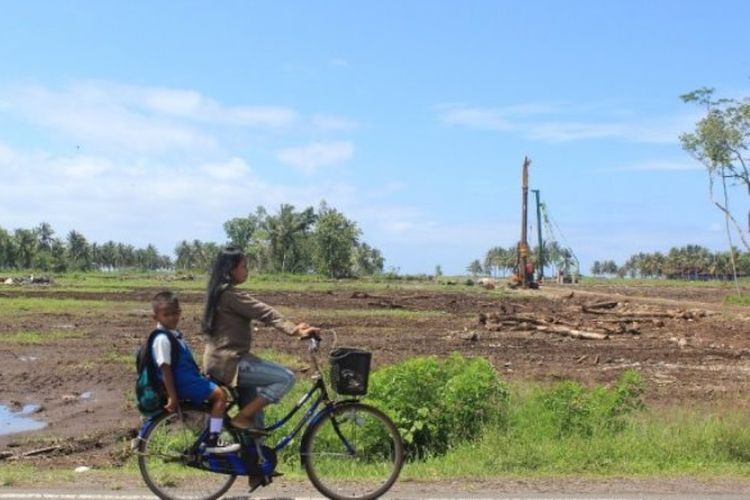 Warga melintas dengan sepeda kayuh di dekat lahan proyek pembangunan bandara New Yogyakarta International Airport di Temon, Selasa (14/11/2017). Tampak di latar belakang, aktivitas pekerja mengoperasikan instalasi pemasang tiang pancang atau paku bumi dalam proyek tersebut. 