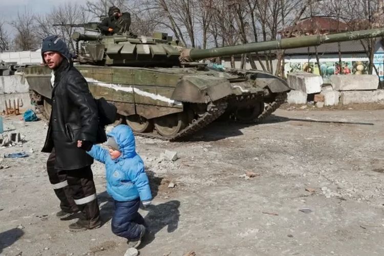 Seorang warga lokal berjalan dengan seorang anak melewati tank pasukan pro-Rusia di kota Mariupol yang sudah terkepung (18/3/2022).