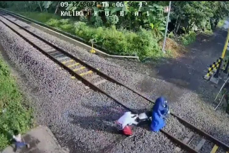 tangkapan layar seorang perempuan bersama dua anak saat mencoba menarik sepeda motor yang mati di tengah rel kereta api 