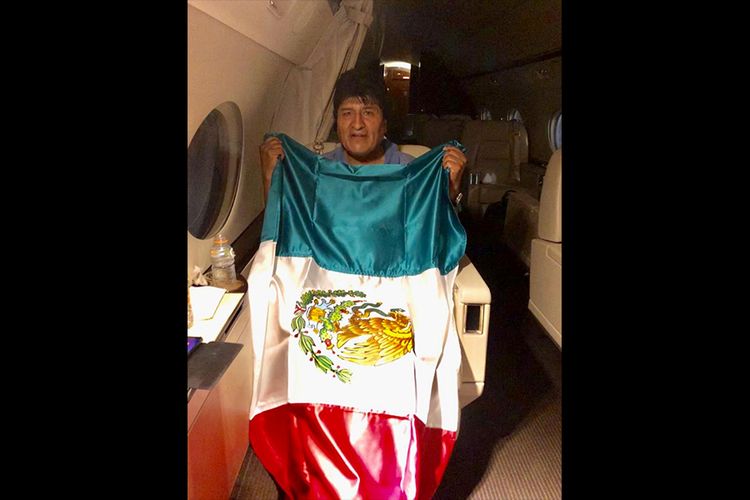 Eks Presiden Bolivia Evo Morales membentangkan bendera negara di tengah pesawat yang akan membawanya ke Meksiko, di mana dia mendapatkan suaka politik pasca-mengundurkan diri pada Minggu (10/11/2019).
