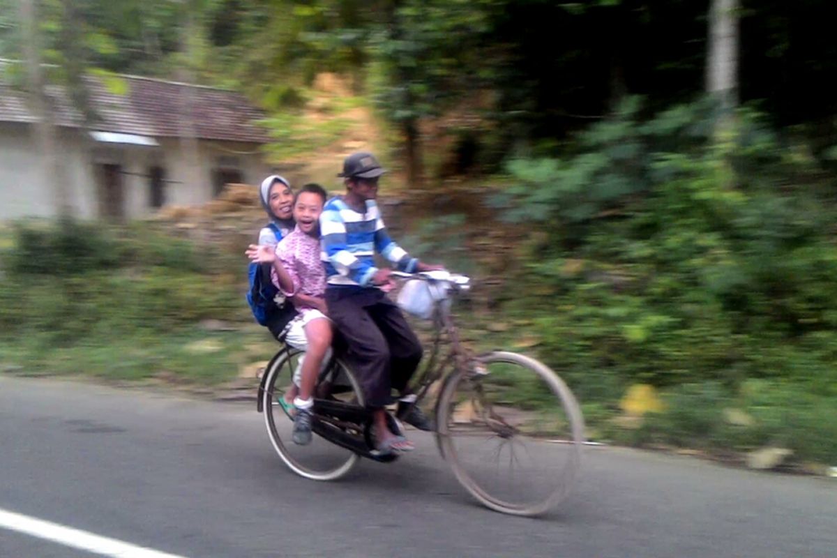Hernowo mengendalikan kemudi sepeda, Kamilah dan Wahyu di belakang. Seperti inilah setiap hari Hernowo membawa Wahyu sekolah yang jauhnya belasan kilometer. Hernowo yang setengah tuli sejak lahir tidak menyerah menyekolahkan anaknya yang down syndrome. Di usia senja mereka, ia mengharapkan Wahyu bisa cepat mandiri.