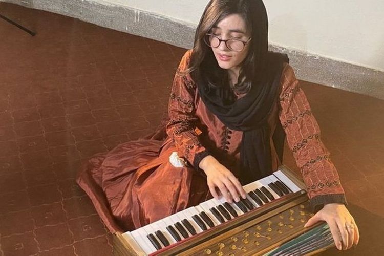 Ayesha Khan duduk di atas selembar karpet coklat, bernyanyi diiringi suara harmonium.
