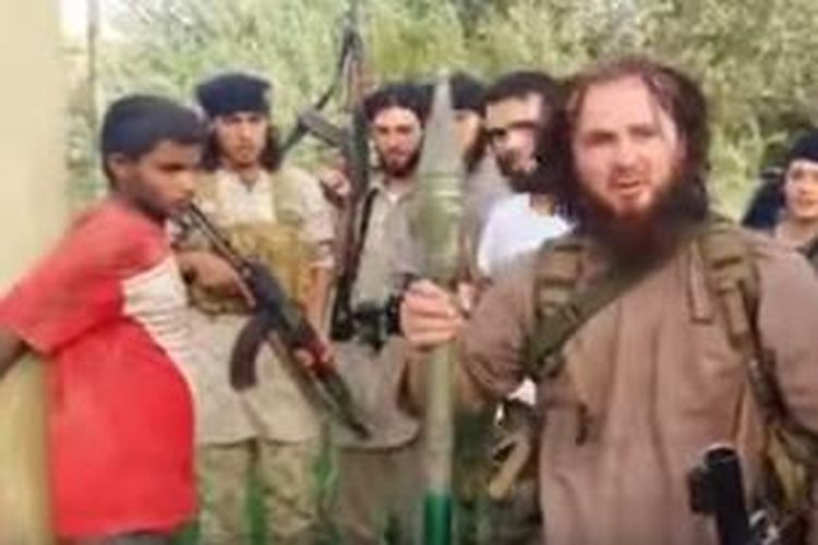 Foto yang diambil dari sebuah video yang diunggah ke internet menampilkan seorang anggota ISIS tengah memegang sebuah peluru bazoka atau RPG. Sementara, si tawanan (baju merah) sudah diikat di sebuah tiang.