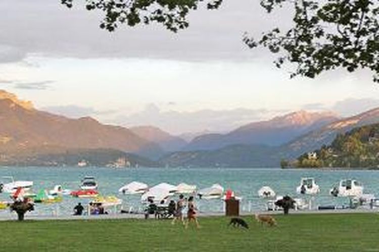 Danau Annecy di Perancis saat sore hari dipenuhi aktivitas berjalan-jalan atau menuntun hewan peliharaan dengan pemandangan matahari tenggelam. Danau ini disebut sebagai danau terbersih di Eropa. 