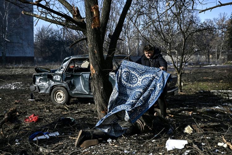Seorang pria menggunakan karpet untuk menutupi tubuh yang tergeletak di tanah setelah pemboman dari serangan udara Rusia di kota Chuguiv, wilayah Kharkiv, Ukraina timur, Kamis (24/2/2022).
