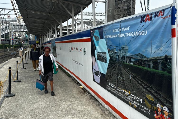 Penumpang Commuter Line melintas di area Stasiun Bogor dengan latar baliho permohonan maaf terkait pekerjaan revitalisasi peron dan penyesuaian layanan perjalanan.
