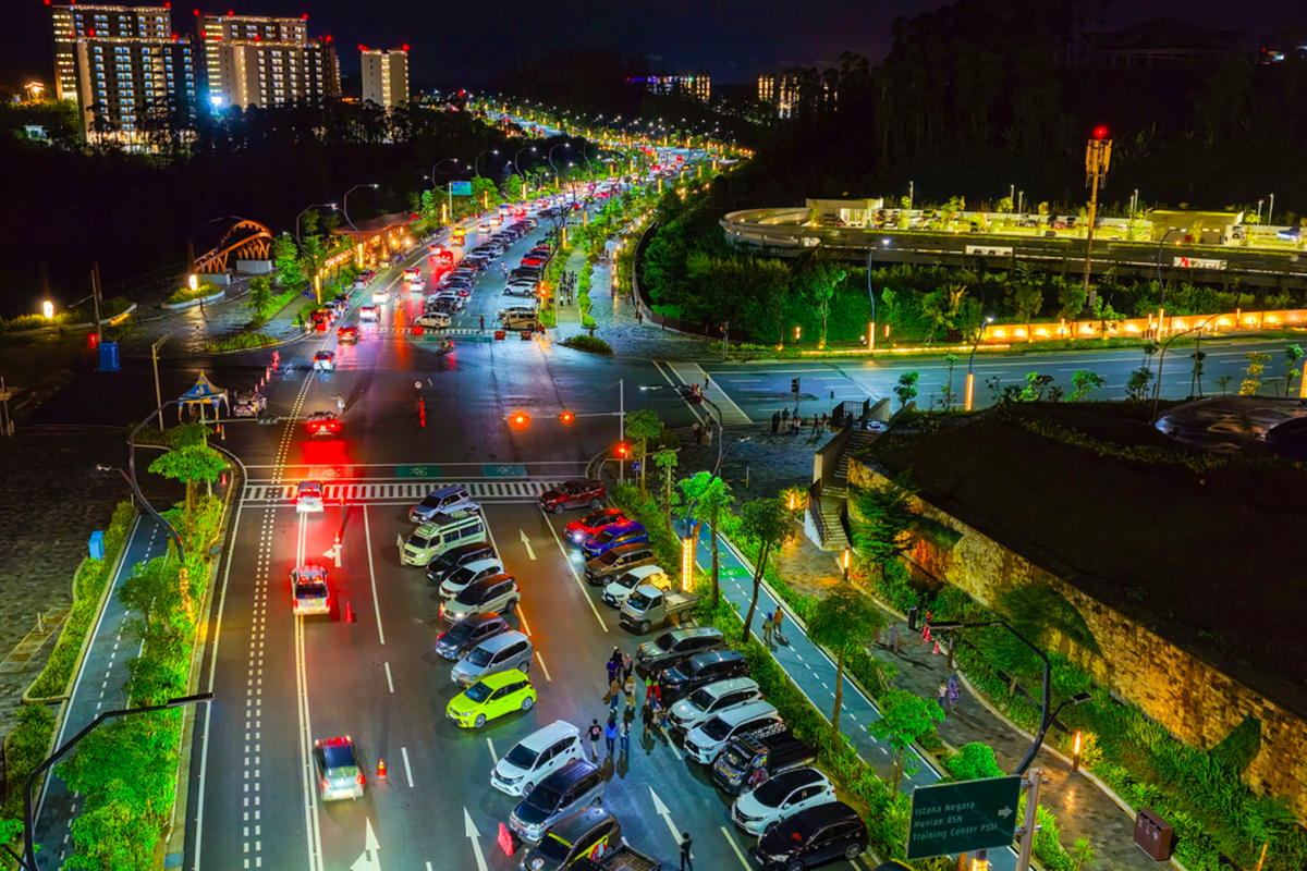 Dari udara, terlihat garis-garis lampu kendaraan yang tertib mengalir di antara gedung-gedung yang berpendar kuning keemasan.
