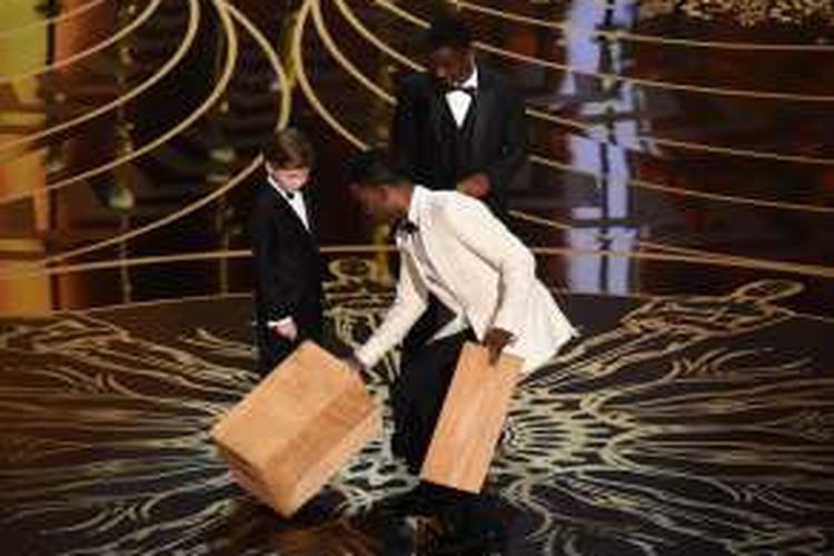 Aktor Chris Rock membawa kotak-kotak kayu agar aktor cilik Jacob Tremblay (kiri) dan Abraham Attah (kanan) bisa mencapai mikrofon di panggung Academy Awards ke-88 di Dolby Theatre, Hollywood, California, Minggu (28/2/2016).