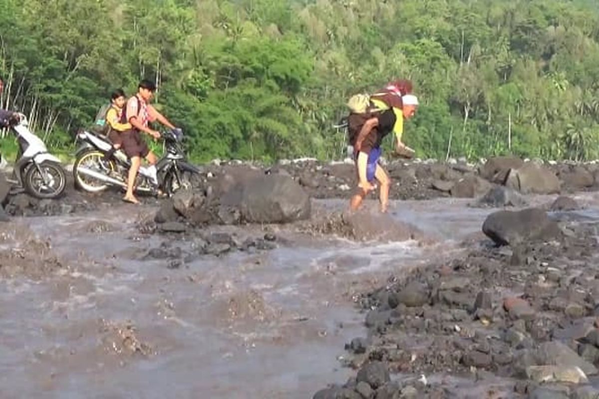 Orang tua siswa di Desa Jugosari menggendong anaknya berangkat ke sekolah menyebrangi aliran lahar Gunung Semeru, Jumat (15/11/2024)