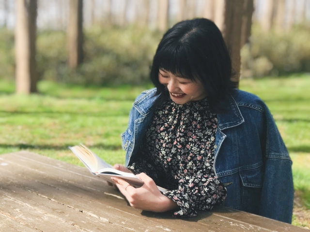 Ilustrasi perempuan memakai blus dan outer sedang membaca buku di taman.